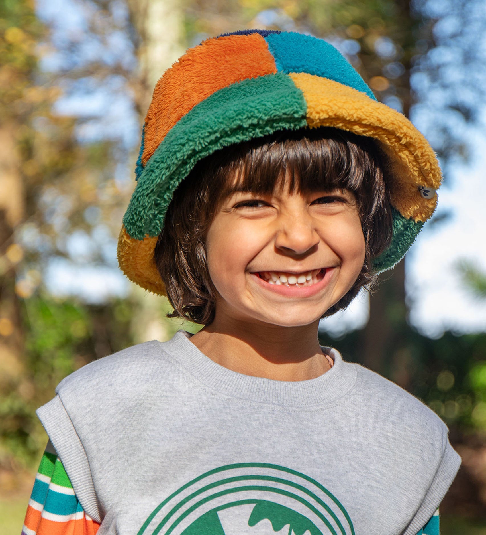 A child standing outside and wearing the Frugi kids fleece bucket hat in hotchpotch. A 100% recycled polyester thick fleeced bucket hat with a multicoloured block design