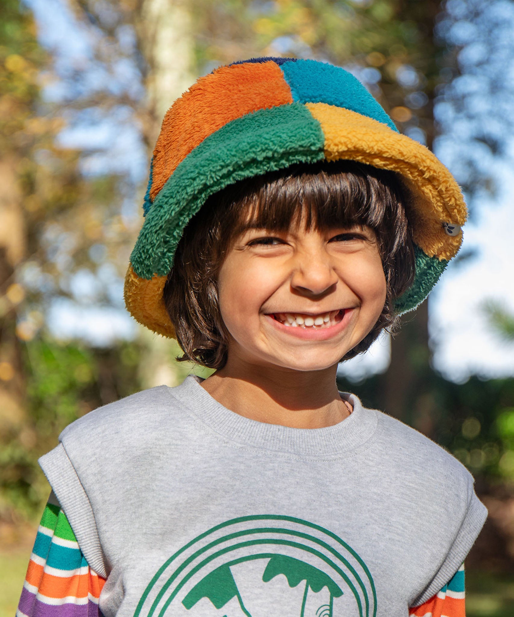 A child standing outside and wearing the Frugi kids fleece bucket hat in hotchpotch. A 100% recycled polyester thick fleeced bucket hat with a multicoloured block design