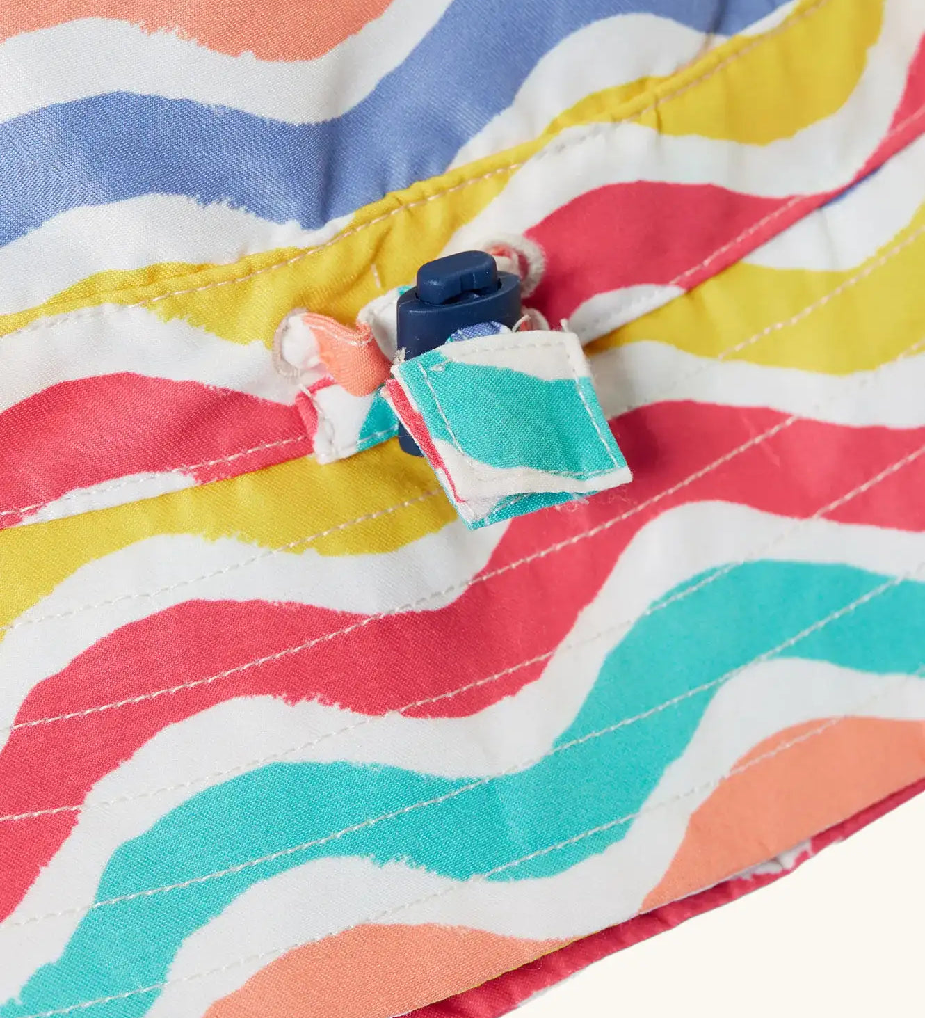 Close up of the toggle on the Frugi adjustable swim hat with a rainbow wave print in pink, white, yellow, purple, peach and turquoise.