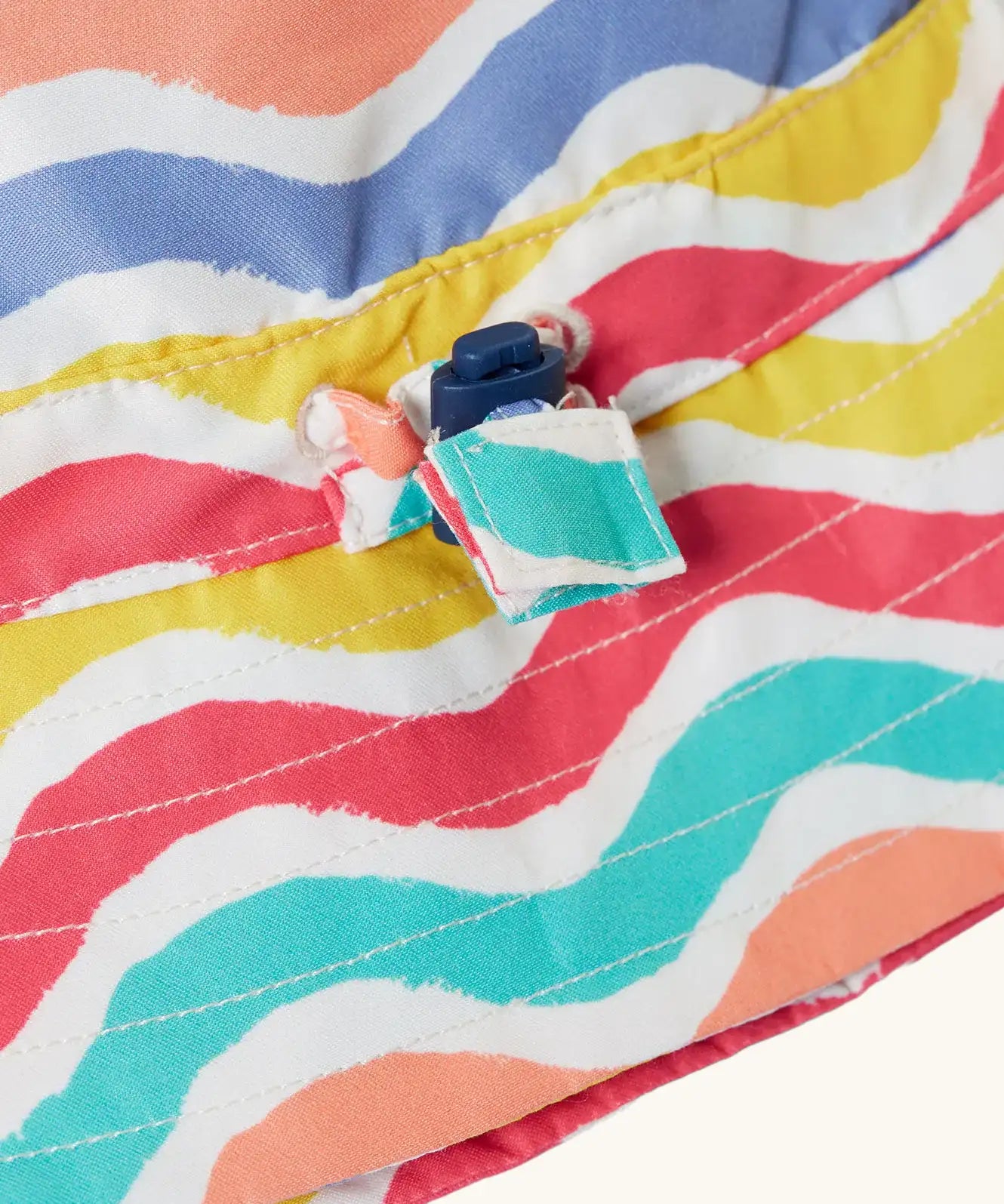 Close up of the toggle on the Frugi adjustable swim hat with a rainbow wave print in pink, white, yellow, purple, peach and turquoise.