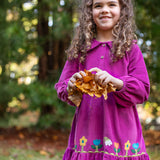 A child holding a bunch of leaves and wearing the frugi holly dress in damson flowers. A bright purple organic cotton dress in a corduroy style with pretty flower appliques on the skirt. 
