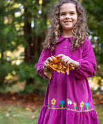 A child holding a bunch of leaves and wearing the frugi holly dress in damson flowers. A bright purple organic cotton dress in a corduroy style with pretty flower appliques on the skirt. 
