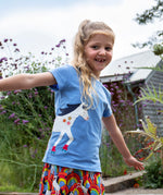 A child playing outside wearing the Frugi Elise wrap around t-shirt . A cornflower blue coloured t-shirt with with short sleeves. Features an appliqué of a white horse wrapped around the front and back of the t-shirt.