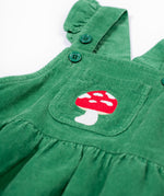 A close up of the frugi kids isla dugaree dress in holly green/mushroom showing the mushroom embroidery detail on the small front pocket and corduroy detail on green organic cotton fabric.