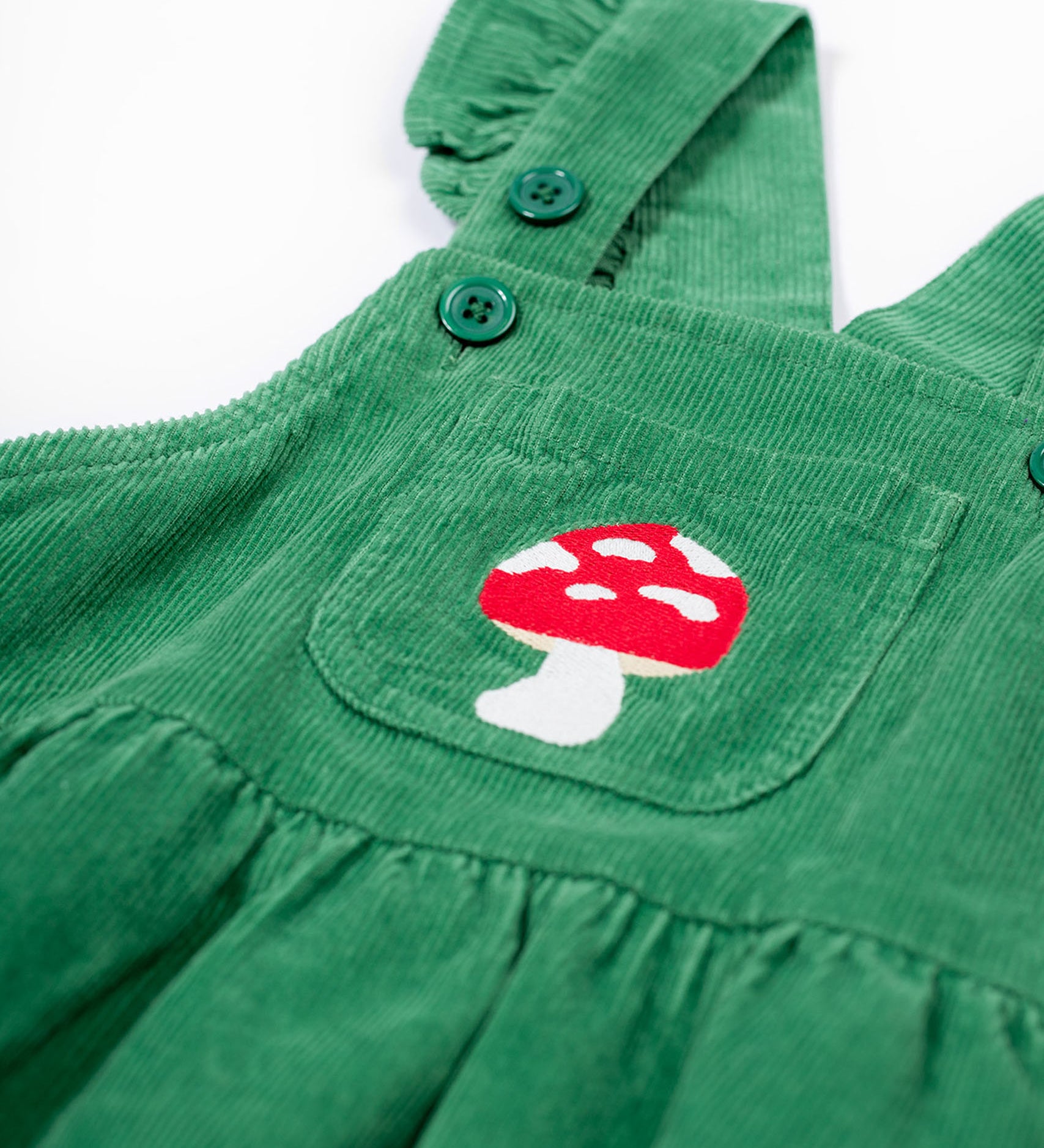 A close up of the frugi kids isla dugaree dress in holly green/mushroom showing the mushroom embroidery detail on the small front pocket and corduroy detail on green organic cotton fabric.