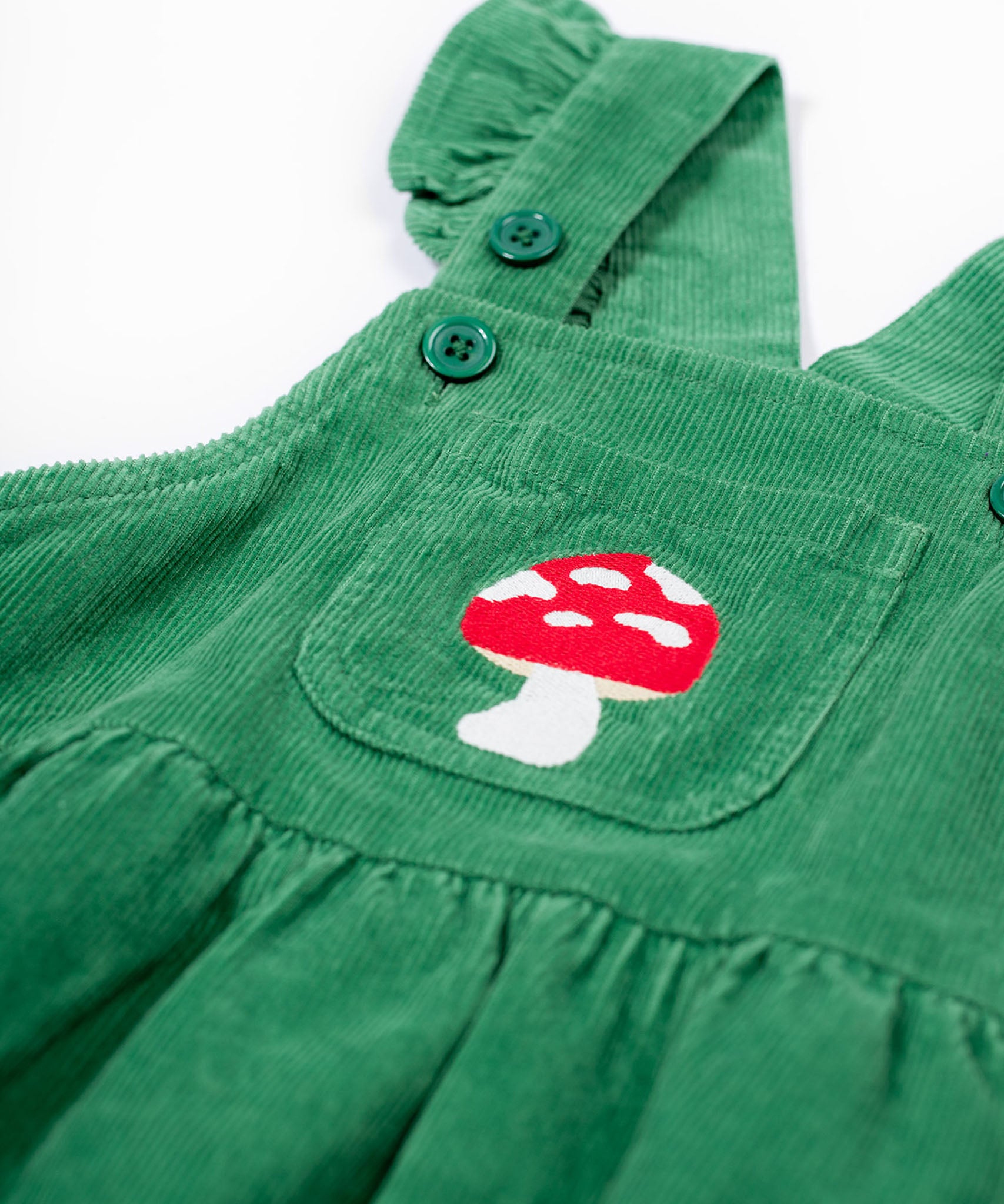 A close up of the frugi kids isla dugaree dress in holly green/mushroom showing the mushroom embroidery detail on the small front pocket and corduroy detail on green organic cotton fabric.