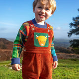 Child wearing the Frugi sebby brown rust organic cotton cord dungaree with a green chest pocket featuring a highland cow applique with a knitted jumper underneath.