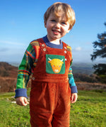 Child wearing the Frugi sebby brown rust organic cotton cord dungaree with a green chest pocket featuring a highland cow applique with a knitted jumper underneath.