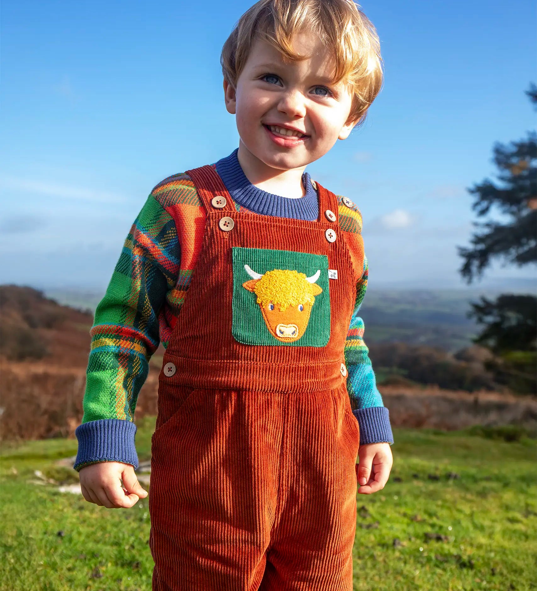 Child wearing the Frugi sebby brown rust organic cotton cord dungaree with a green chest pocket featuring a highland cow applique with a knitted jumper underneath.