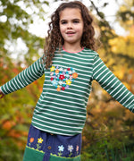 A little girl in the forest wearing an organic cotton Frugi outfit - the Cara Corduroy Skirt with flower embroidery, and a long sleeved embroidered stripy top.