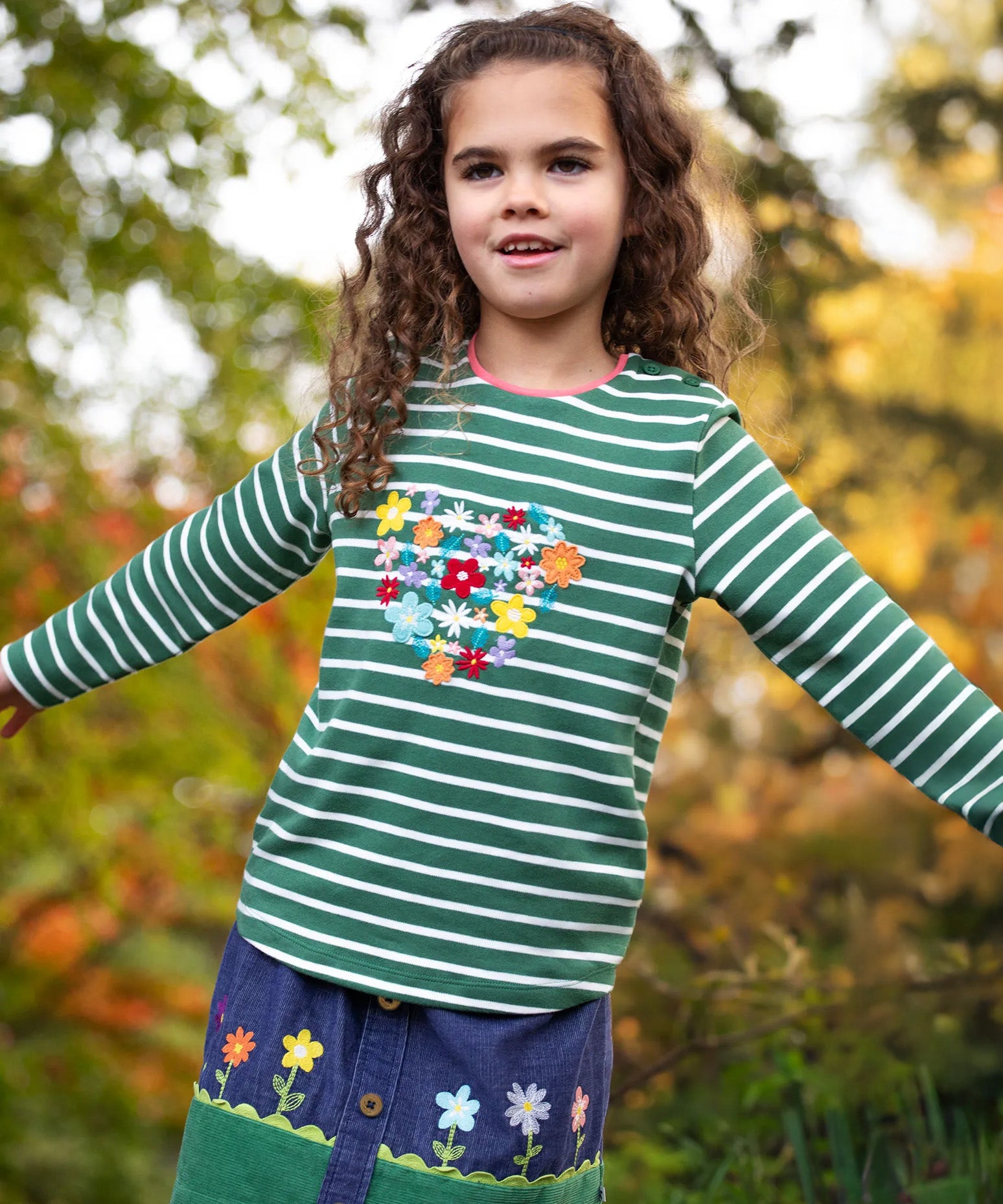 A little girl in the forest wearing an organic cotton Frugi outfit - the Cara Corduroy Skirt with flower embroidery, and a long sleeved embroidered stripy top.