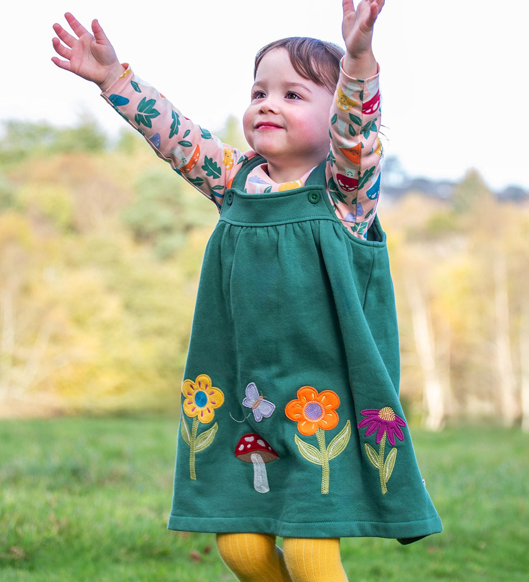 A child standing outside and wearing the Frugi kids pippa holly green fun guys pinafore outfit. A 100% GOTS organic cotton woodland inspired outfit which features a pink long sleeve top and gree n pinafore