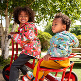 Two children of a red pedal trike both wearing Frugi waterproof puddle buster coats. One in pink purrfect ponies print and one in the blue terrific tractors print.
