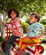 Two children of a red pedal trike both wearing Frugi waterproof puddle buster coats. One in pink purrfect ponies print and one in the blue terrific tractors print.