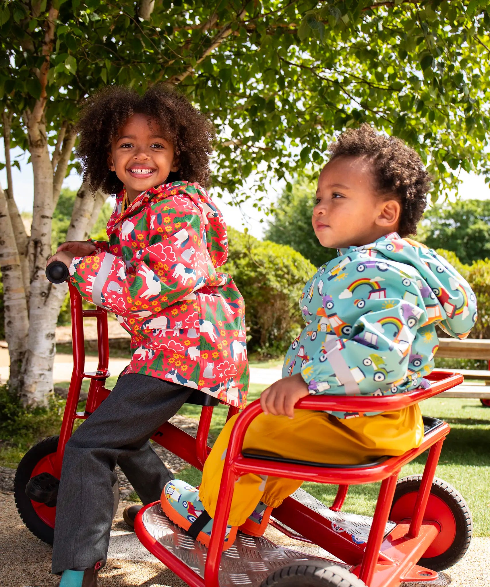 Two children of a red pedal trike both wearing Frugi waterproof puddle buster coats. One in pink purrfect ponies print and one in the blue terrific tractors print.