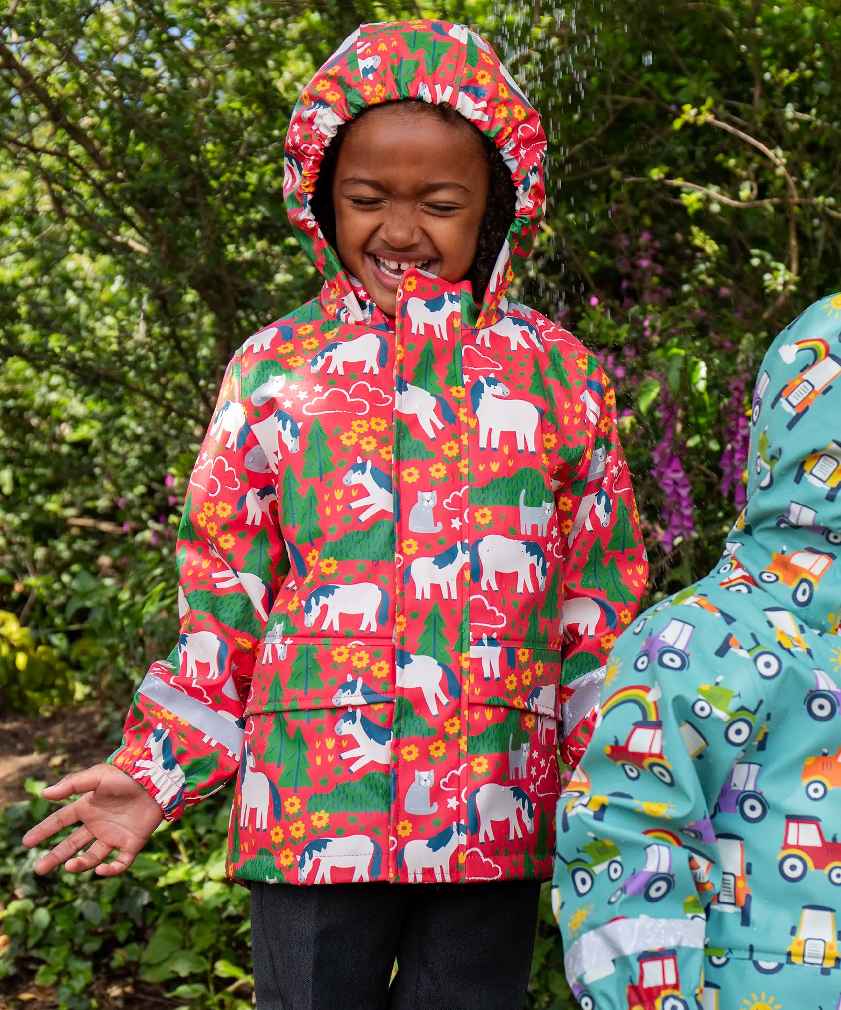 Child wearing Frugi waterproof puddle buster coat in pink purrfect ponies print with the hood up.
