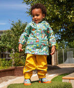Child wearing Frugi waterproof puddle buster coat in blue terrific tractors  print with yellow puddle buster trousers and matching terrific tractor wellington boots. 