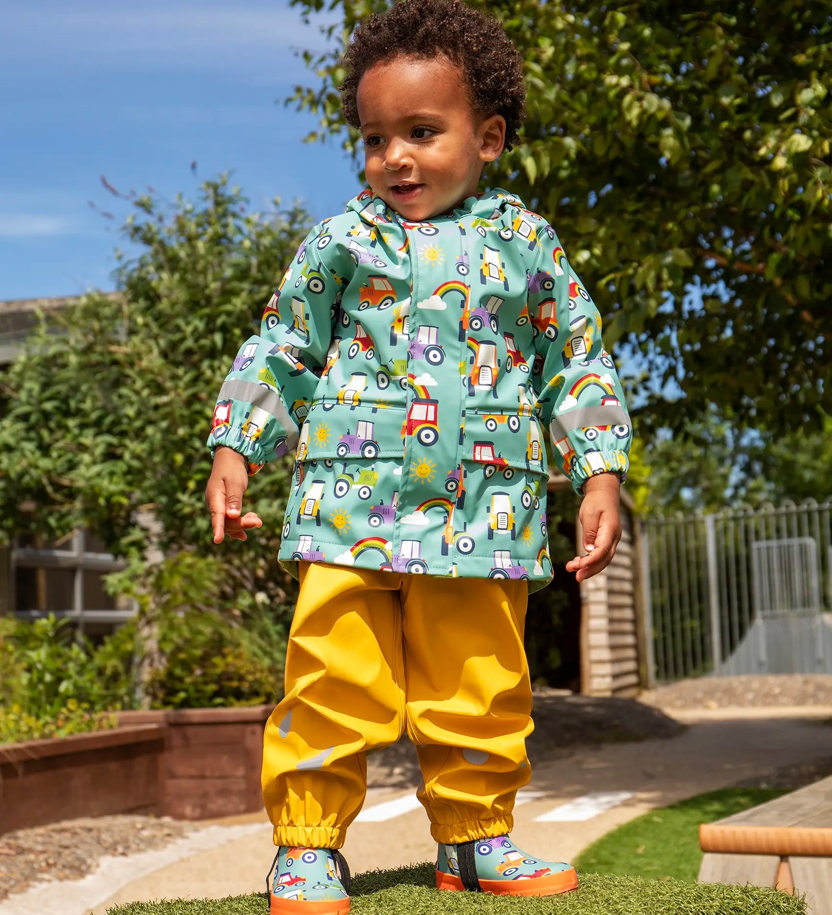 Child wearing Frugi waterproof puddle buster coat in blue terrific tractors  print with yellow puddle buster trousers and matching terrific tractor wellington boots. 