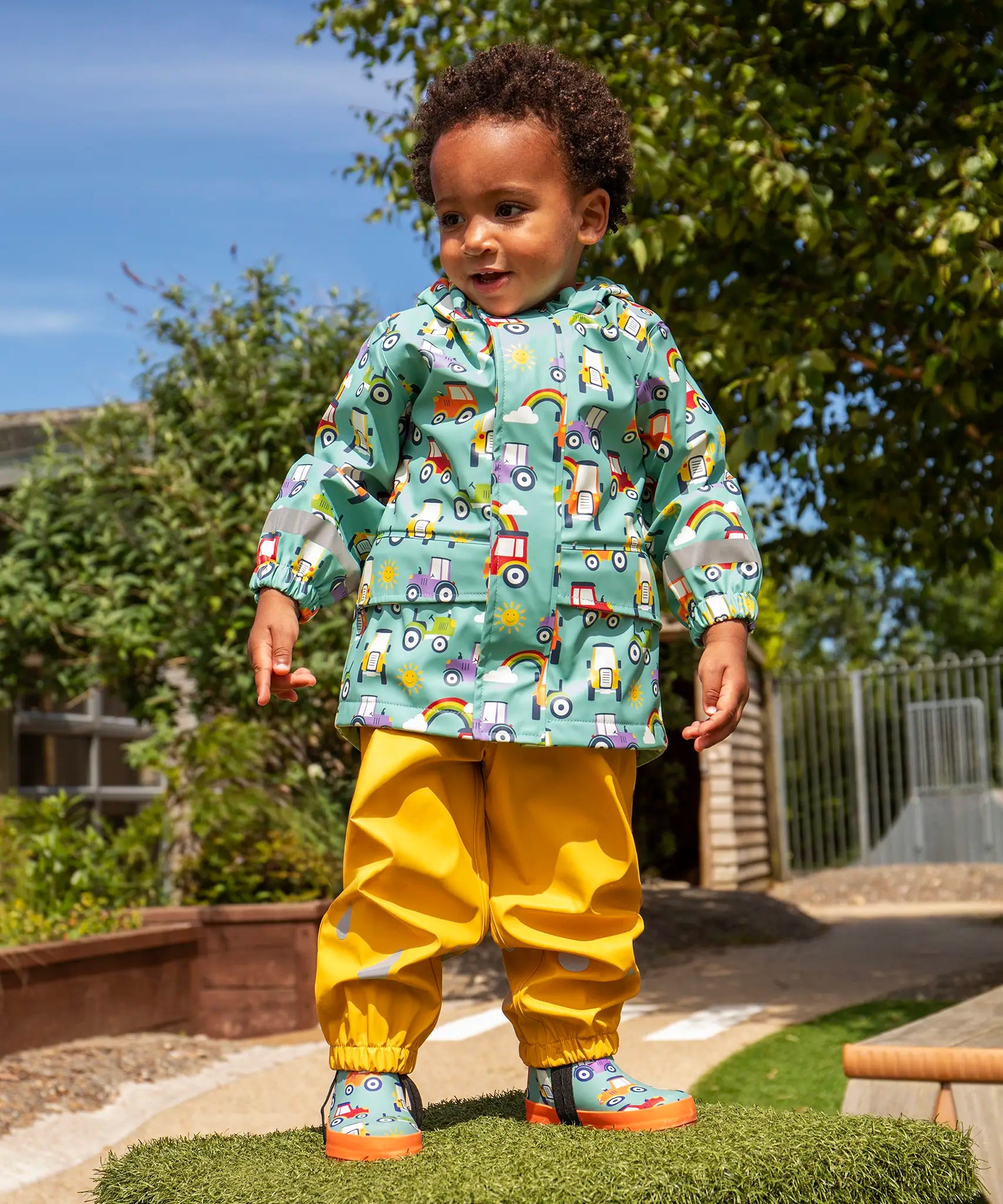 Child wearing Frugi waterproof puddle buster coat in blue terrific tractors  print with yellow puddle buster trousers and matching terrific tractor wellington boots. 