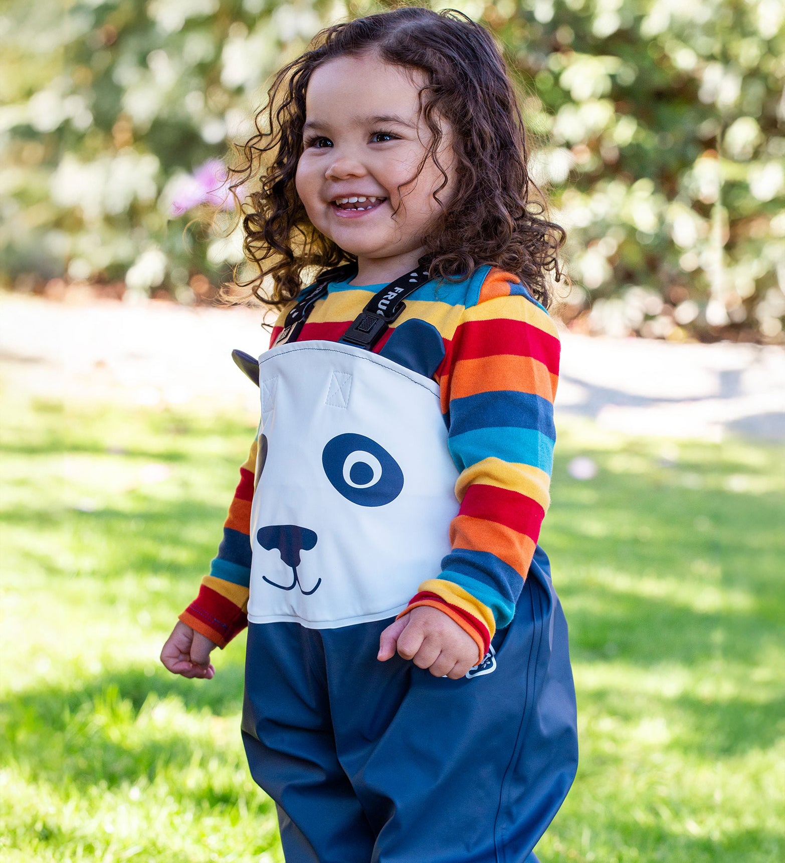 A child wearing the Frugi puddle buster trousers in navy / panda over a rainbow long sleeve top. A waterproof trouser with shoulder straps for a secure fit. 
