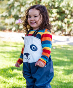 A child wearing the Frugi puddle buster trousers in navy / panda over a rainbow long sleeve top. A waterproof trouser with shoulder straps for a secure fit. 