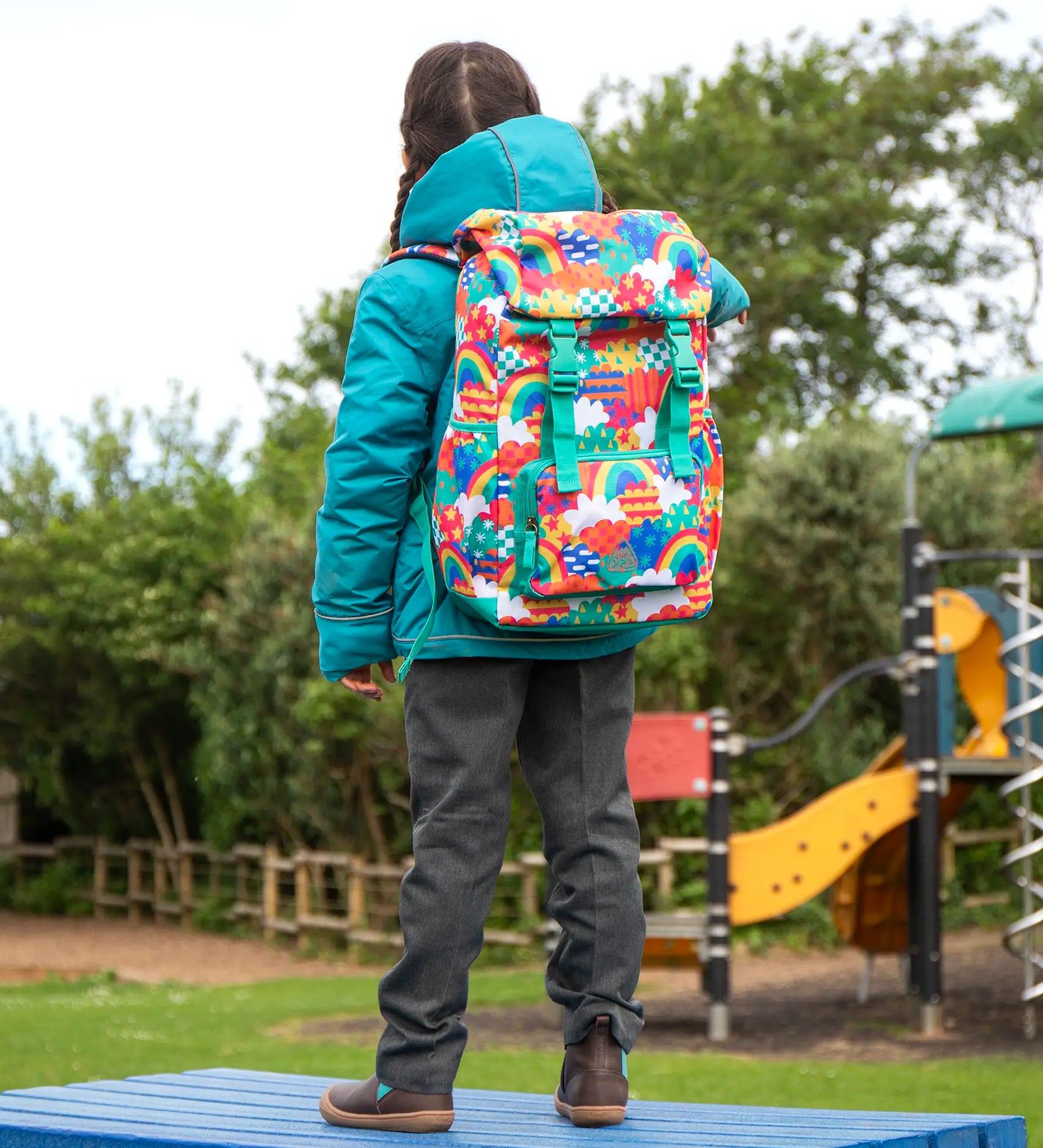 View of the back of a child wearing the Frugi kids 3 in 1 rambler coat in boat blue rhubarb check with a colourful Frugi Trail Blazing fun frenzy backpack on her back.