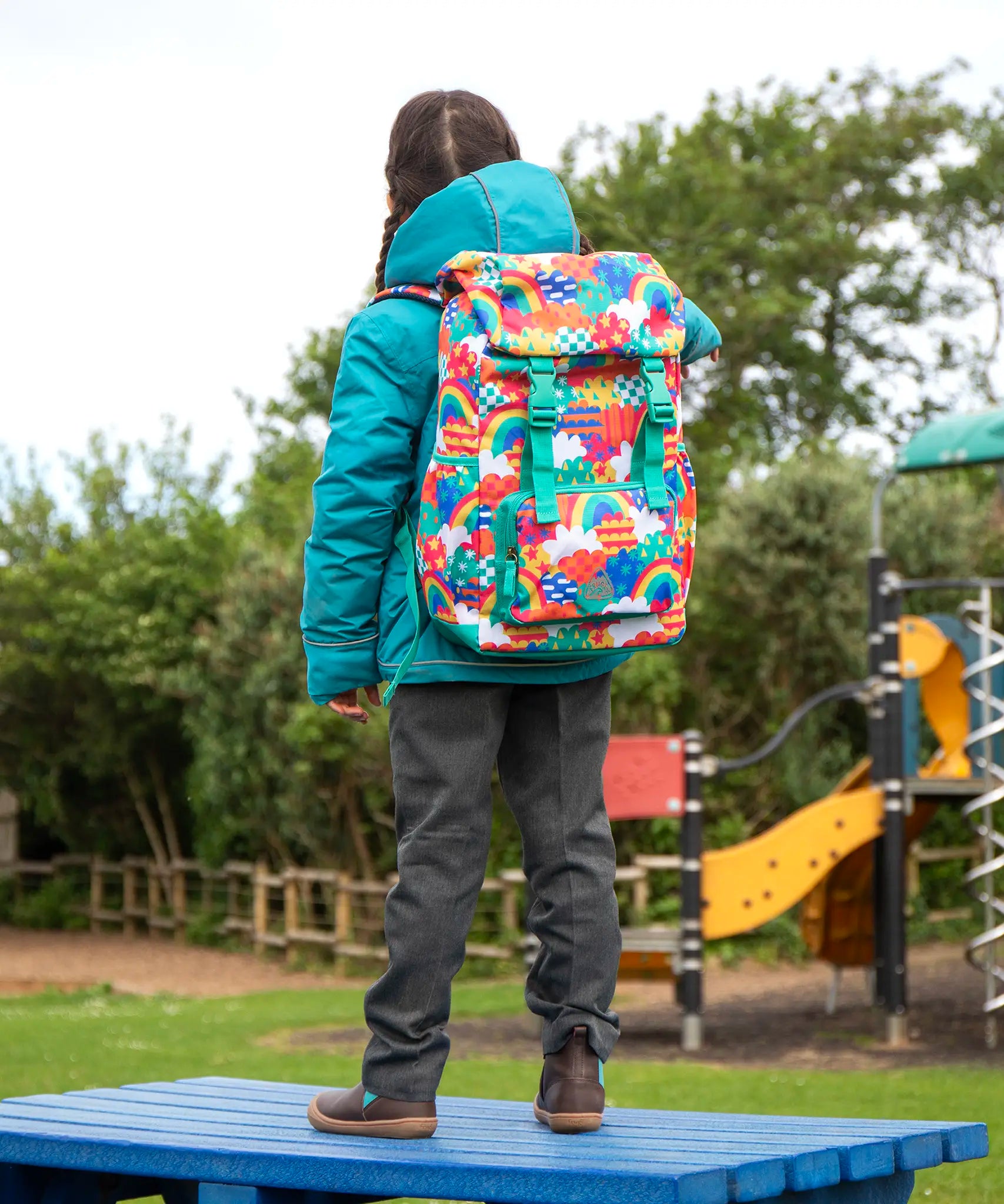 View of the back of a child wearing the Frugi kids 3 in 1 rambler coat in boat blue rhubarb check with a colourful Frugi Trail Blazing fun frenzy backpack on her back.