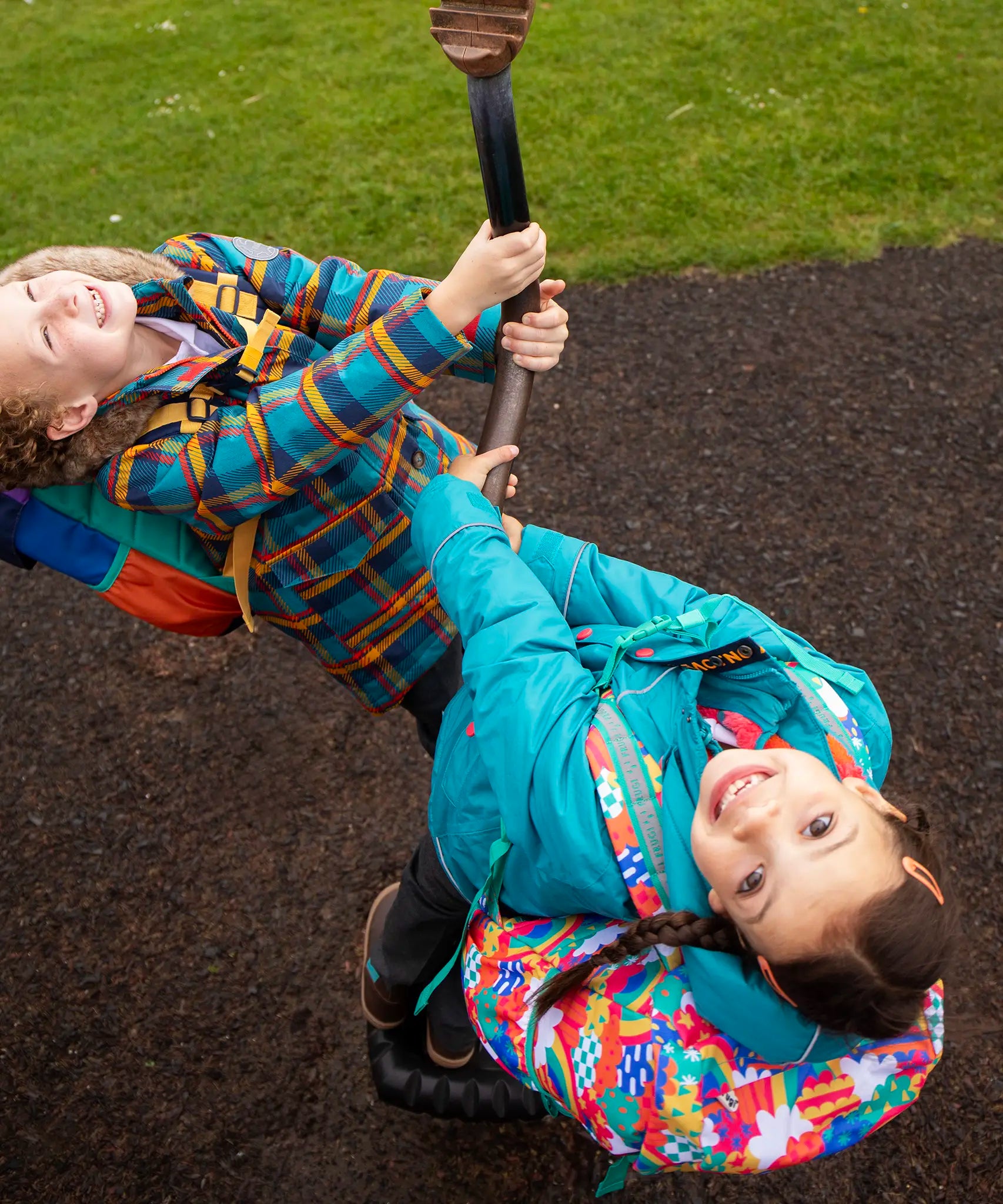 Two children in a playground wearing Frugi coats. One is wearing the Frugi kids 3 in 1 rambler coat in boat blue rhubarb.