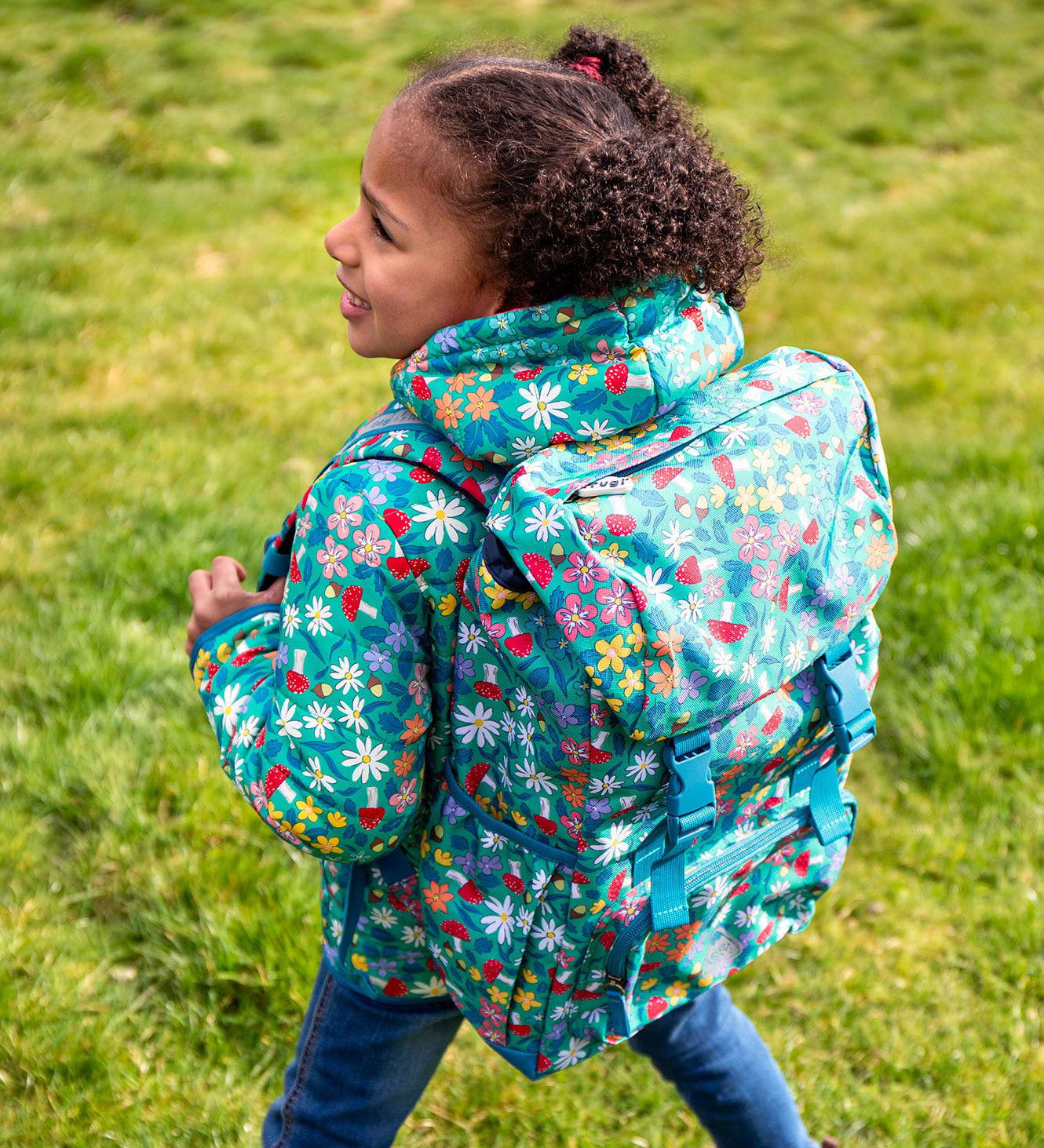 A child standing outside with a Frugi trailblazing backpack in wildwoods backpack on the back. A Light green backpack with woodland inspired prints all over. Features 2 buckle fasteners on the lid, a small zipped compartment on the front and 2 bottle holders.