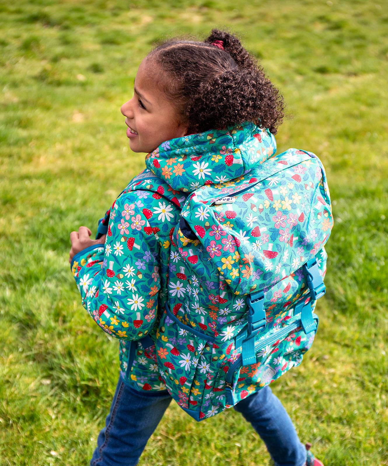 A child standing outside with a Frugi trailblazing backpack in wildwoods backpack on the back. A Light green backpack with woodland inspired prints all over. Features 2 buckle fasteners on the lid, a small zipped compartment on the front and 2 bottle holders.