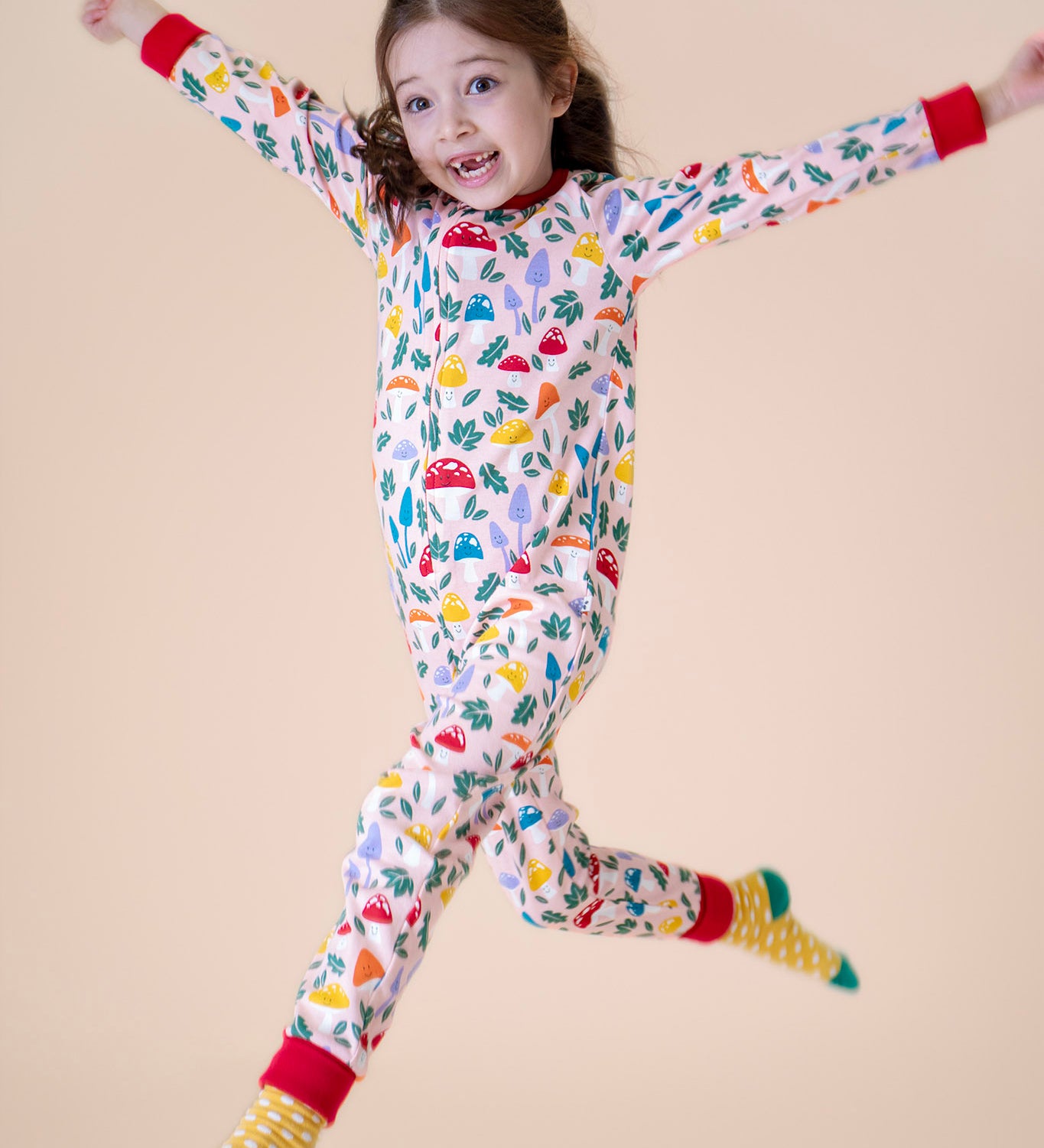 A child jumping in the air and wearing the Frugi Zelah fun guys all in one zip suit available at Babipur. A GOTS organic cotton all in one suit light pink with a playful woodland inspired prints and red cuffs. Features a full length zip.