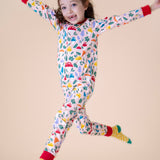 A child jumping in the air and wearing the Frugi Zelah fun guys all in one zip suit available at Babipur. A GOTS organic cotton all in one suit light pink with a playful woodland inspired prints and red cuffs. Features a full length zip.