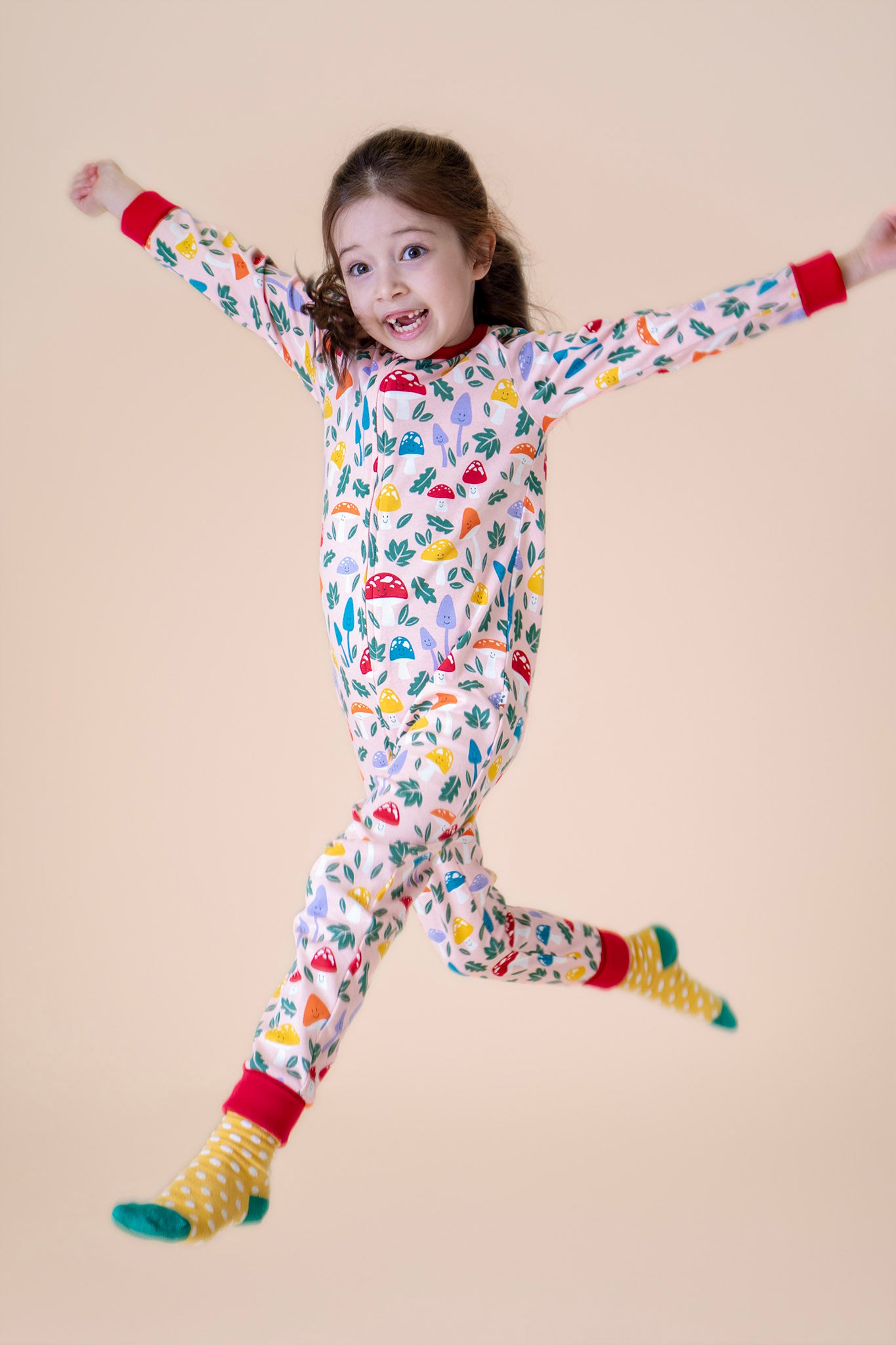 A child jumping in the air and wearing the Frugi Zelah fun guys all in one zip suit available at Babipur. A GOTS organic cotton all in one suit light pink with a playful woodland inspired prints and red cuffs. Features a full length zip.