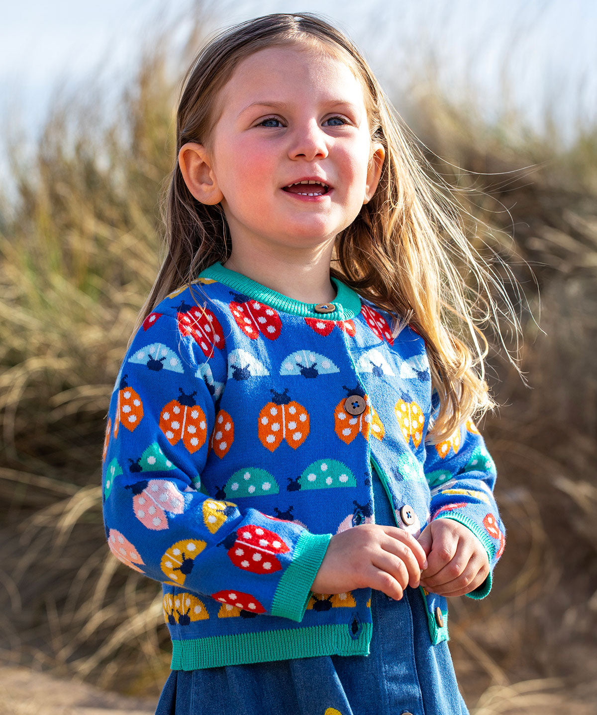 A young girl on sand dunes with long blonde hair wearing a Frugi fair isle style knit cardigan with multicoloured ladybug detail all over with a denim dress underneath. 