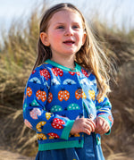 A young girl on sand dunes with long blonde hair wearing a Frugi fair isle style knit cardigan with multicoloured ladybug detail all over with a denim dress underneath. 