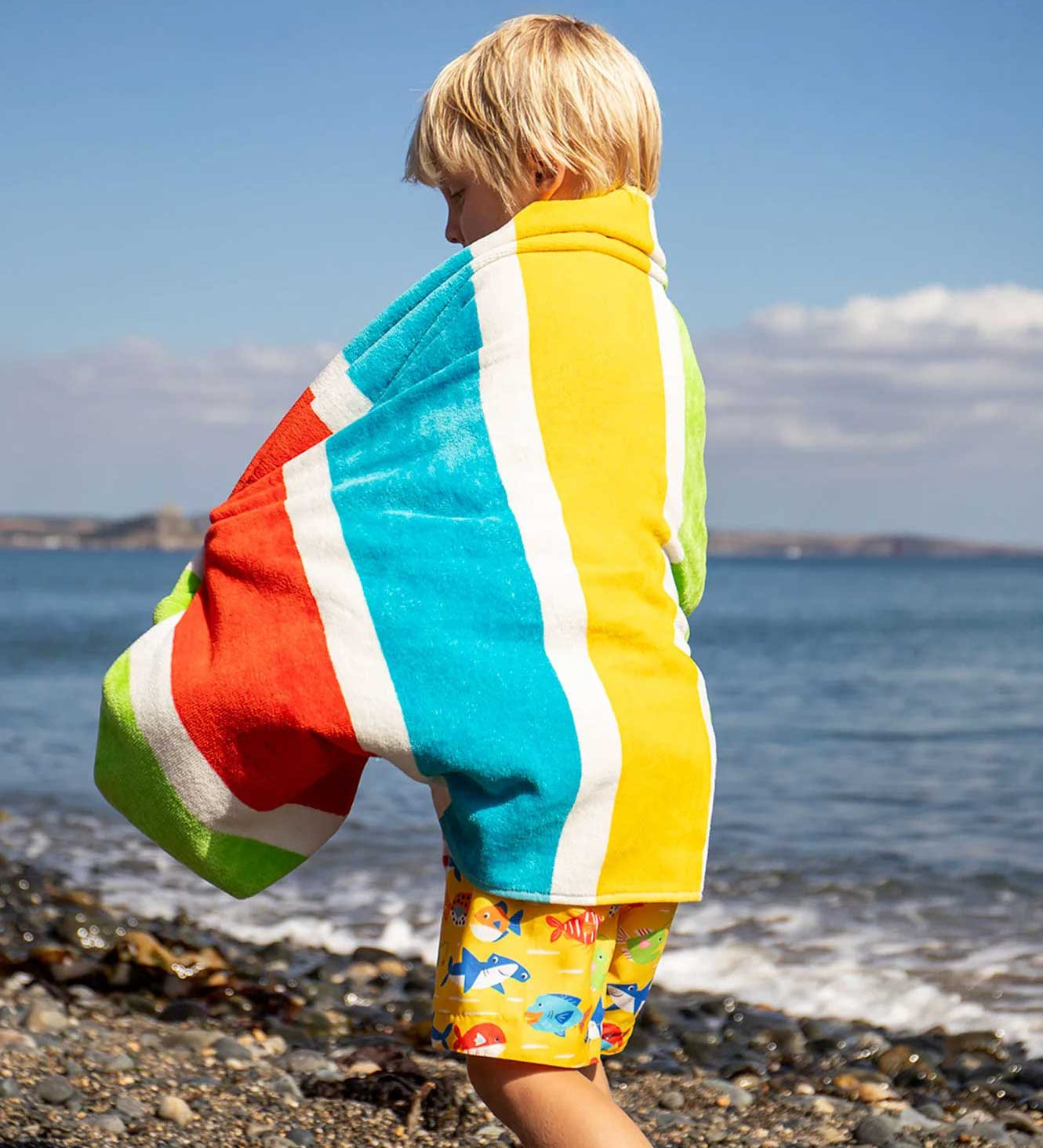 Child wearing the Frugi lagoon organic cotton beach towel with rainbow stripes over their back. 