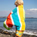 Child wearing the Frugi lagoon organic cotton beach towel with rainbow stripes over their back. 