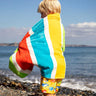 Child wearing the Frugi lagoon organic cotton beach towel with rainbow stripes over their back. 
