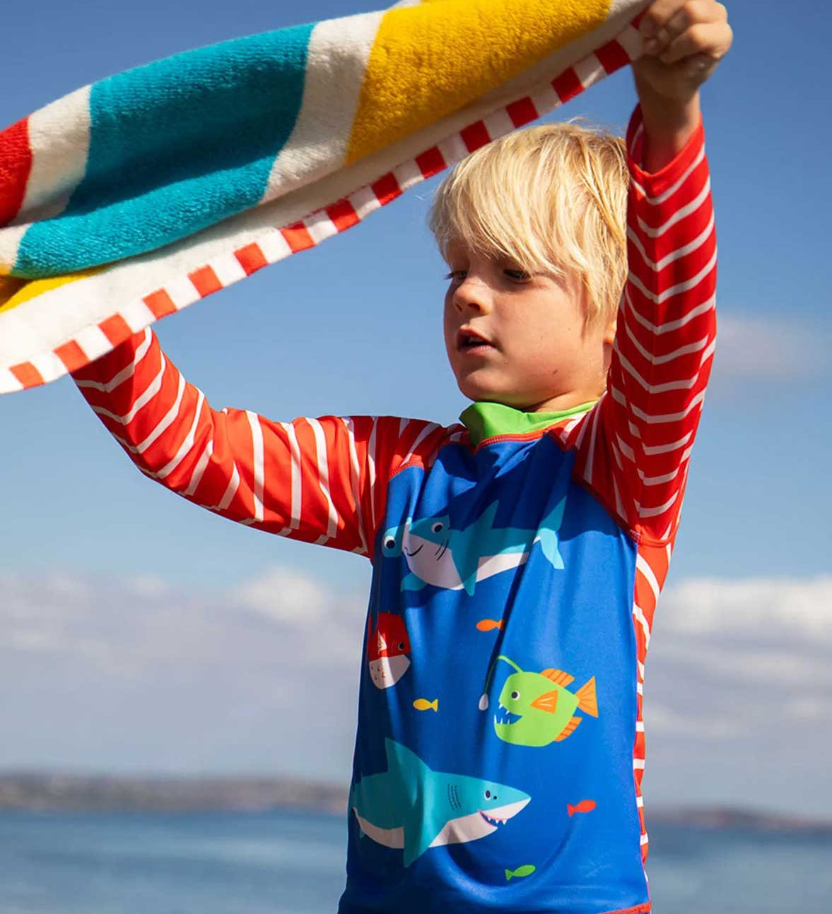A child holding a Frugi rainbow stripe organic cotton beach towel above their head. There is red and white binding on the edges of the towel. 