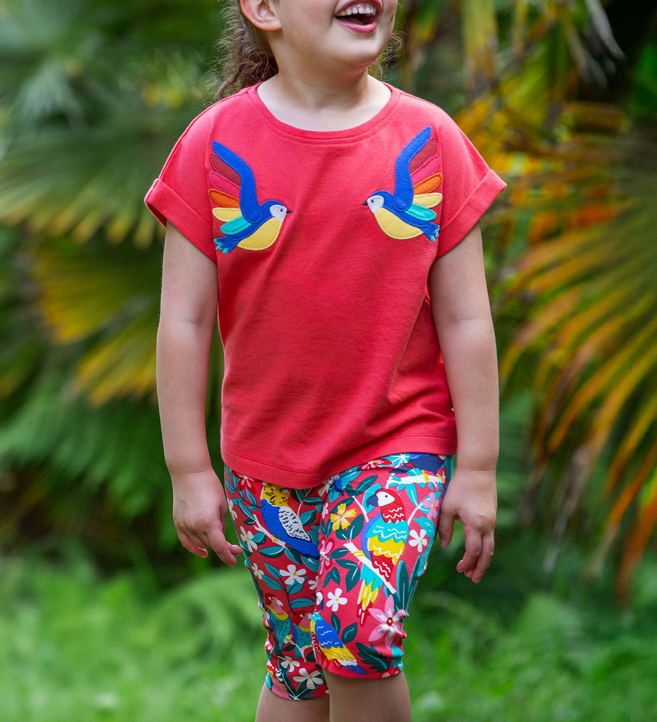 A smiling toddler wearing Frugi Laurie Shorts with a Birds Of Paradise print, featuring different exotic birds surrounded by colourful flowers and leaves on a pink background, walking amongst trees.