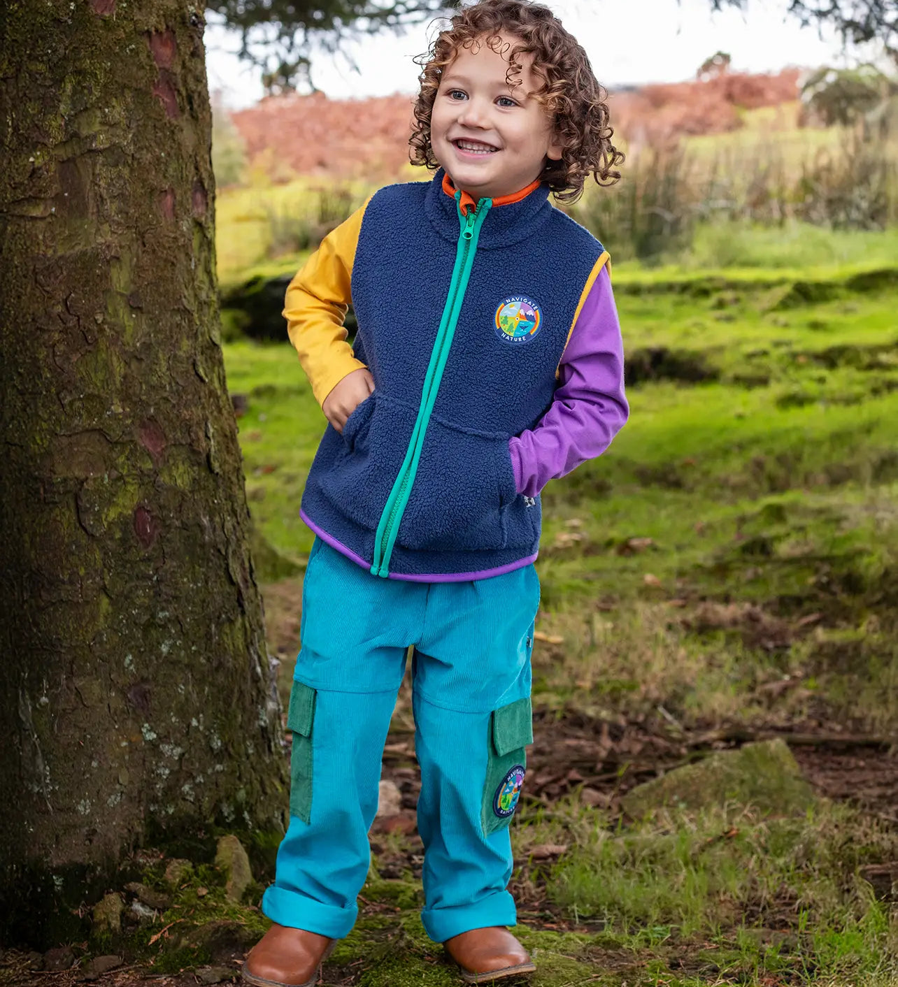 Child wearing the Frugi Lennox adventure trousers in boat blue organic cotton corduroy with a navy fleece gilet and long sleeved raglan top underneath.