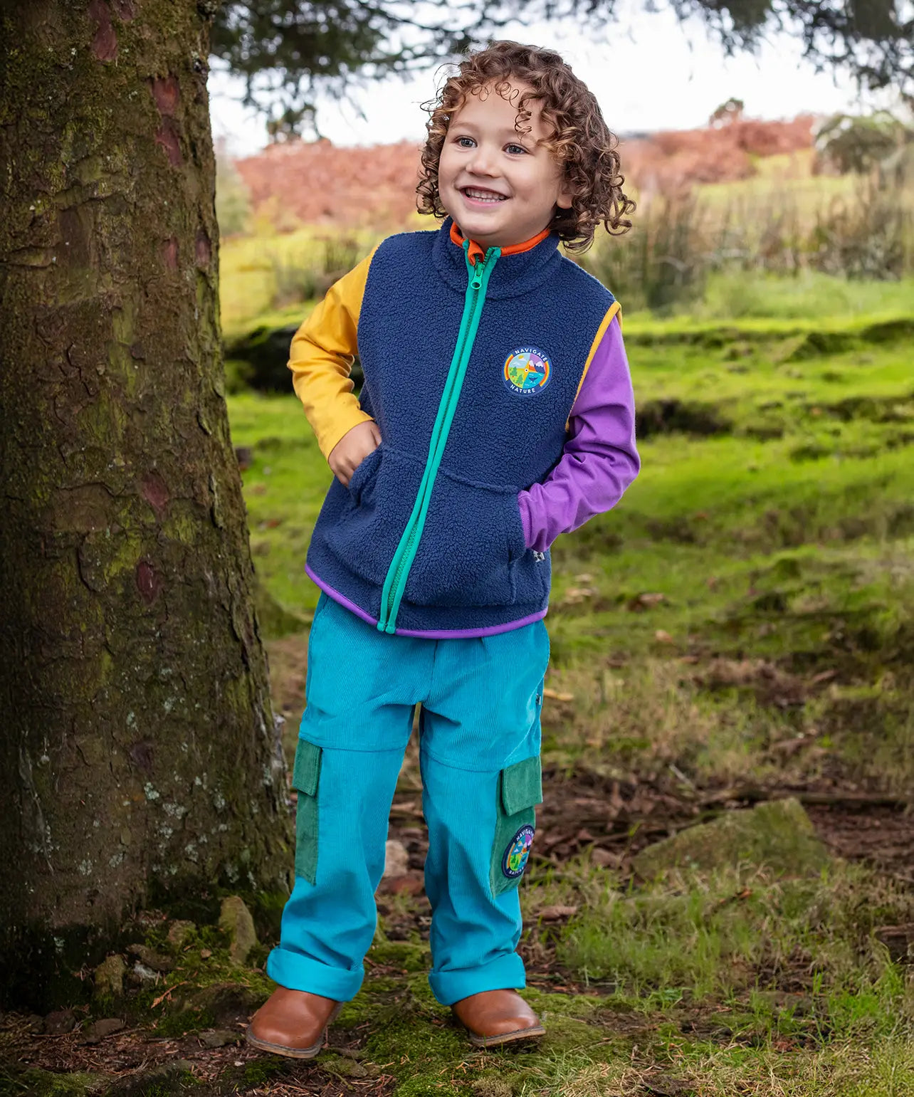 Child wearing the Frugi Lennox adventure trousers in boat blue organic cotton corduroy with a navy fleece gilet and long sleeved raglan top underneath.