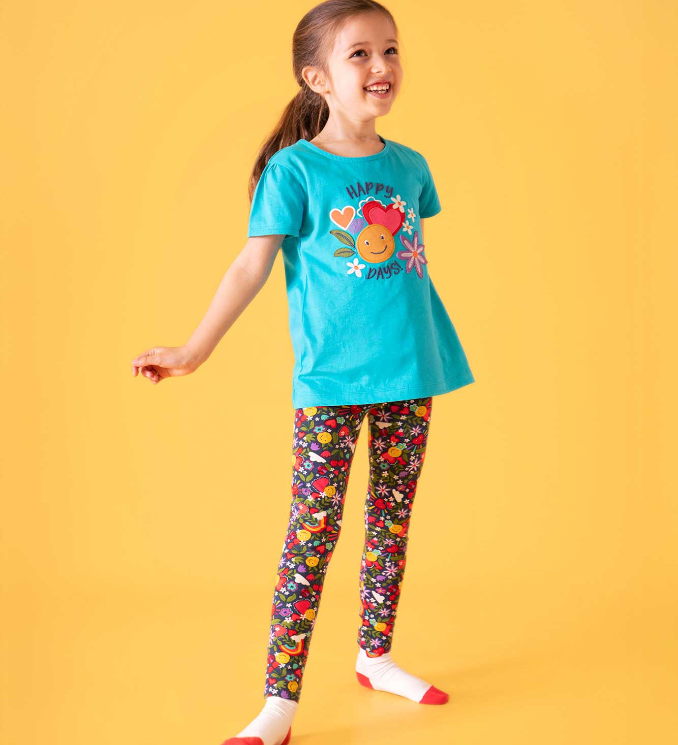 A child with long hair in a ponytail smiling, wearing a blue short sleeves t-short is a happy days graphic on the front and Frugi leggings with an all over happy days print featuring hearts, rainbows, flowers and smiley faces.