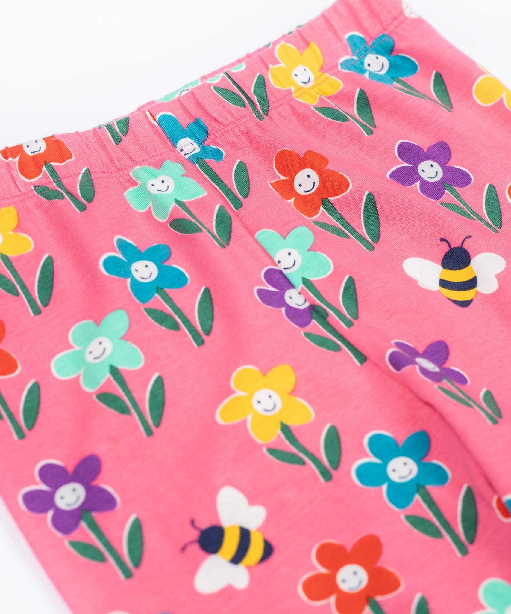 Frugi organic cotton kids Libby leggings with a fun flowers print on a pink background. The flowers are multi-coloured and have happy faces on them.