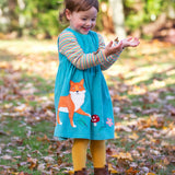 A child playing outdoors wearing a boat blue coloured cord dress from Frugi. The dress has a fox applique on the front just to the side next to a red mushroom and leaves. 