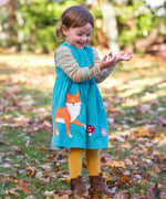 A child playing outdoors wearing a boat blue coloured cord dress from Frugi. The dress has a fox applique on the front just to the side next to a red mushroom and leaves. 