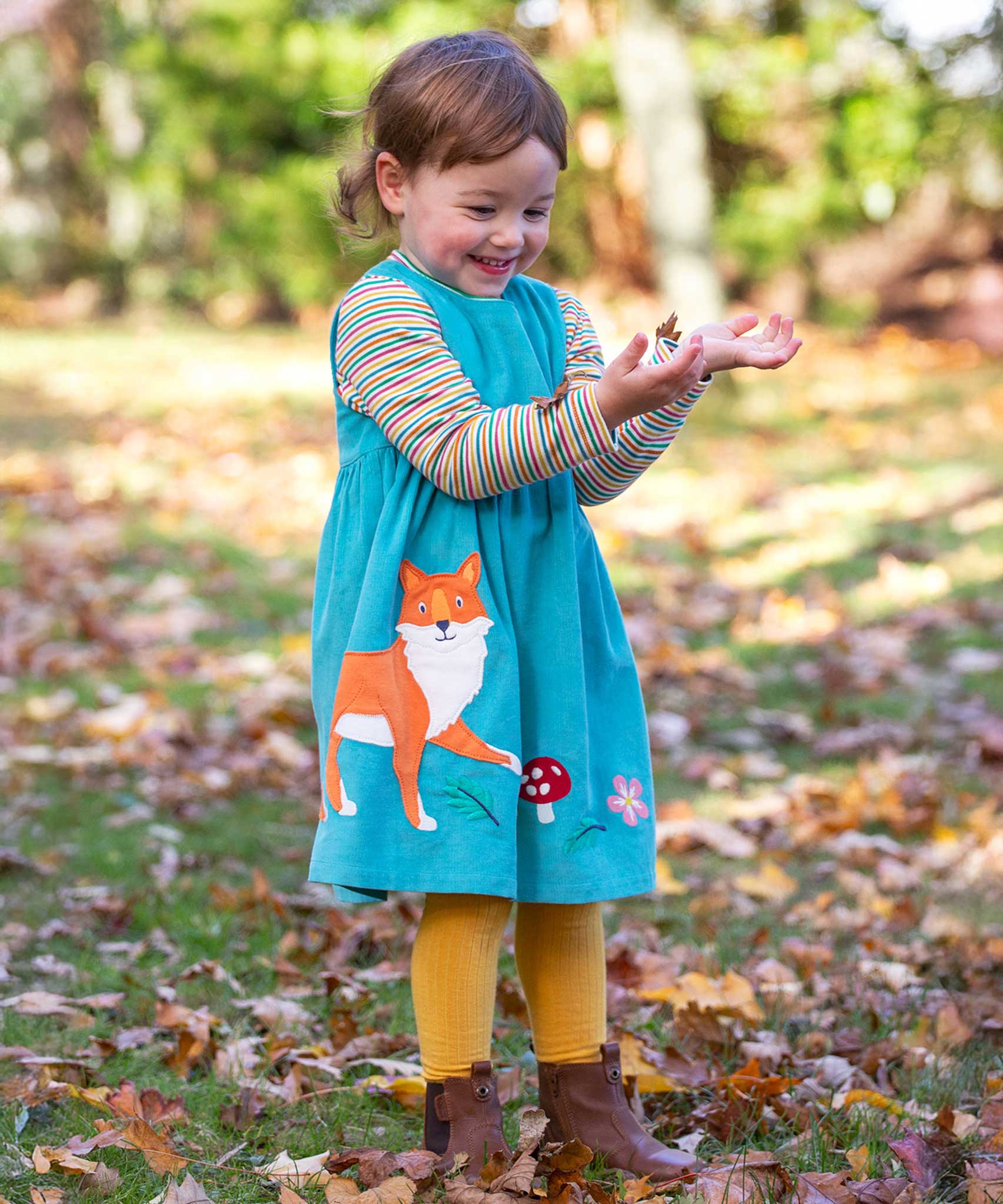 A child playing outdoors wearing a boat blue coloured cord dress from Frugi. The dress has a fox applique on the front just to the side next to a red mushroom and leaves. 