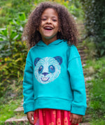 A child standing outside wearing the Frugi oversized Lissie hoody - a light blue colour with a playful panda embroidery on the front. They are wearing the Frugi Samantha Floral Fun / Flamingo Summer Dress underneath the hoody.