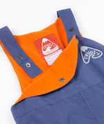 A close up of the Frugi little kids waterproof dungarees in navy showing the popper detail for adjusting the size. Features a orange fleece inner liner for warmth and a name tag stitched on the inside.