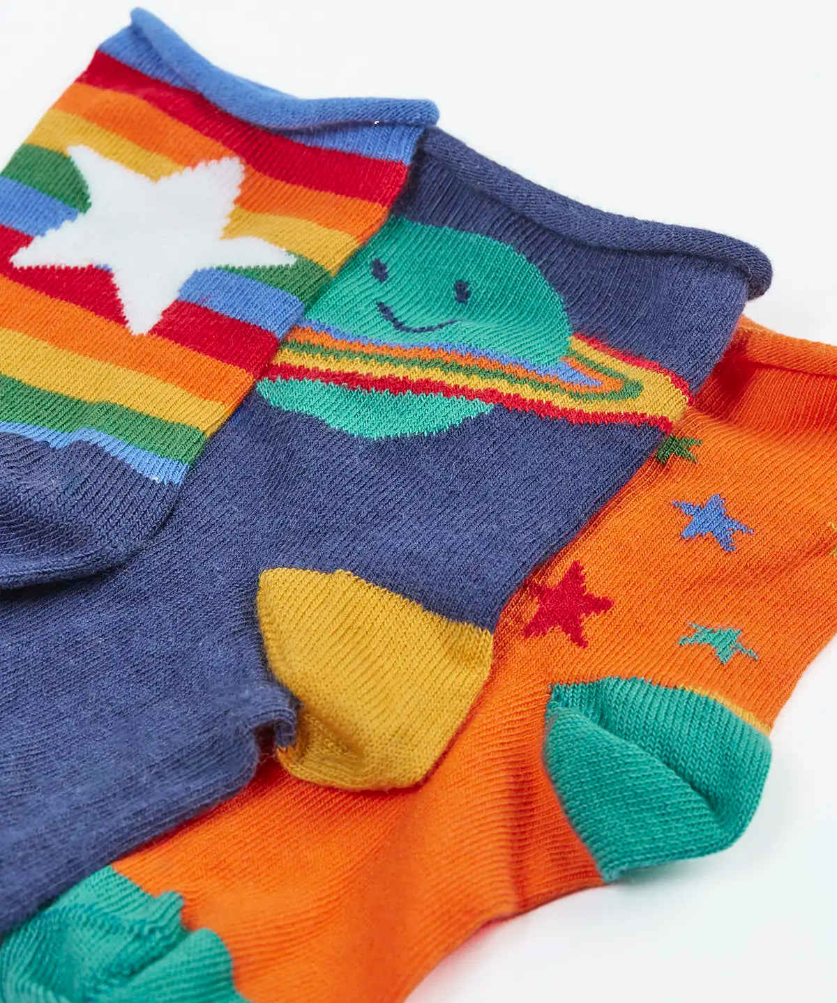Close up of the Frugi little roly bamboo socks in a 3 pack with spac edesign. Rainbow stripe with white star pair, navy with planet pair and an orange with rainbow stars pair.